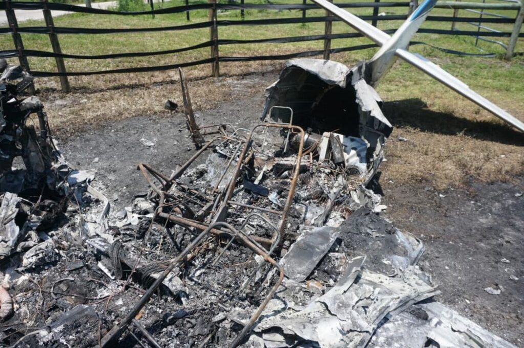 Charred interior fuselage of the Commander 114TC wreckage, with exposed tubular frame and severely burned cockpit area, tail section still attached in background.